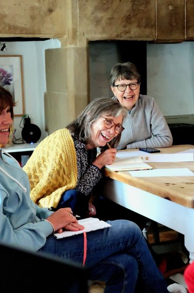 Three smiling women engaged in a discussion around a table in a bright, casual setting.