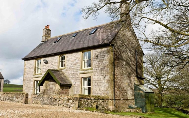 A stone cottage with a sloped roof, surrounded by greenery and rolling hills.