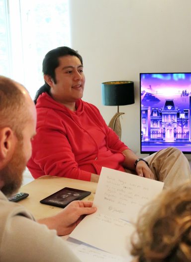 Person in a red hoodie sits at a table discussing notes, with a TV showing a cityscape in the background.