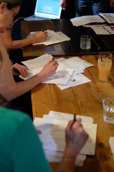 Hands writing on papers at a wooden table, with drinks nearby.