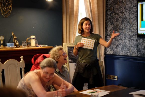 A woman stands presenting to a small group seated at a table in a well-lit room.