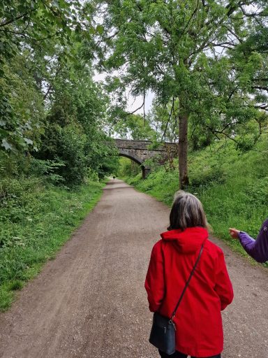 Two people walk along a tree-lined path beside a stone bridge.