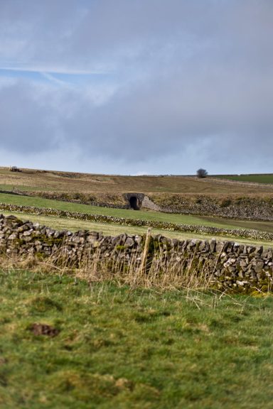 A rural landscape with rolling hills, a stone wall, and a distant bridge under a cloudy sky.