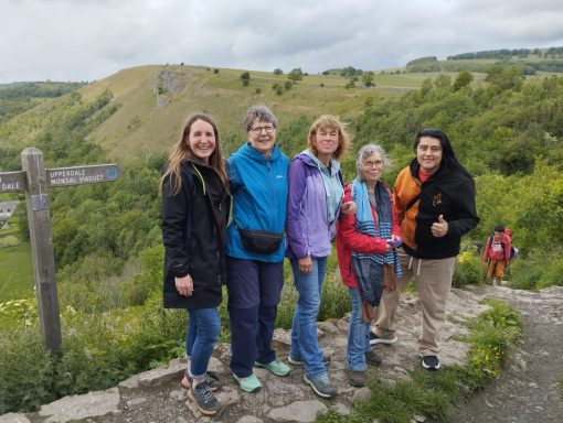 Five people stand on a trail with green hills in the background, smiling and enjoying the scenery.