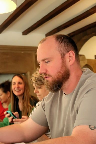 A man with a beard sits at a table, focused on writing, with several people in the background.