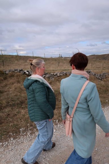 Two women walking on a path in a rural landscape under a cloudy sky.