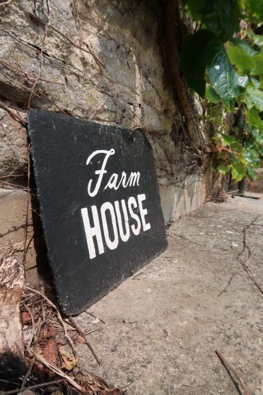 A black slate sign displaying the words "Farm House" leaning against a stone wall.