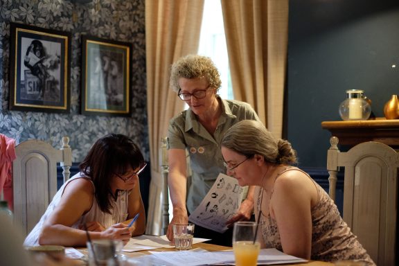 Two women sit at a table, while a third woman stands, discussing papers and notes.