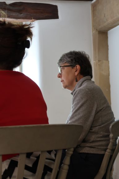 A seated woman with short hair and glasses, facing a window, next to a person in a red top.
