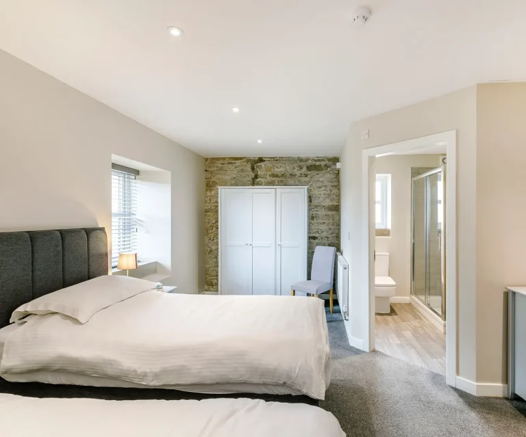 Modern bedroom with two beds, neutral colours, and an ensuite bathroom visible.