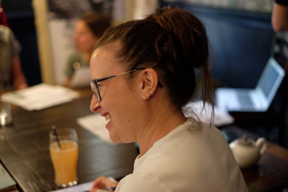 A smiling woman with glasses sitting at a table, wearing a white top, with a drink nearby.