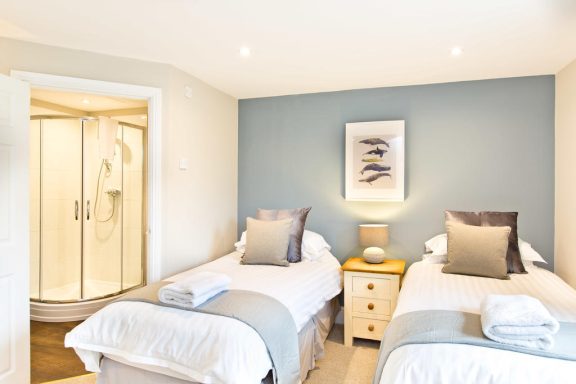 Bright bedroom with two single beds, towels, and an en-suite shower. Light decor and natural light.