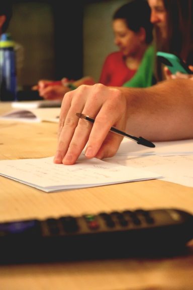 A hand holding a pen over paper with a calculator and people in the background.
