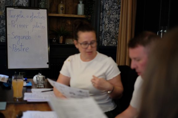 A woman discusses documents with a man at a table, with notes and a drink nearby.
