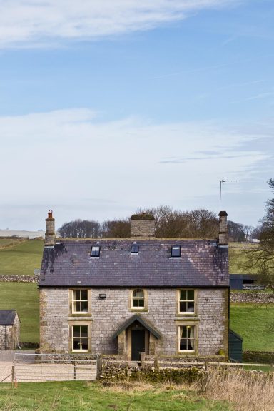 A stone house with a slate roof, set against a clear blue sky and rolling green fields.